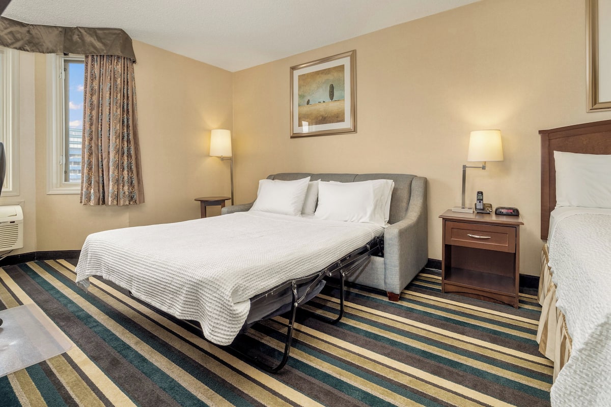 A hotel room with a bed, bedside lamps, a window with curtains, a painting on the wall, and a striped carpet.