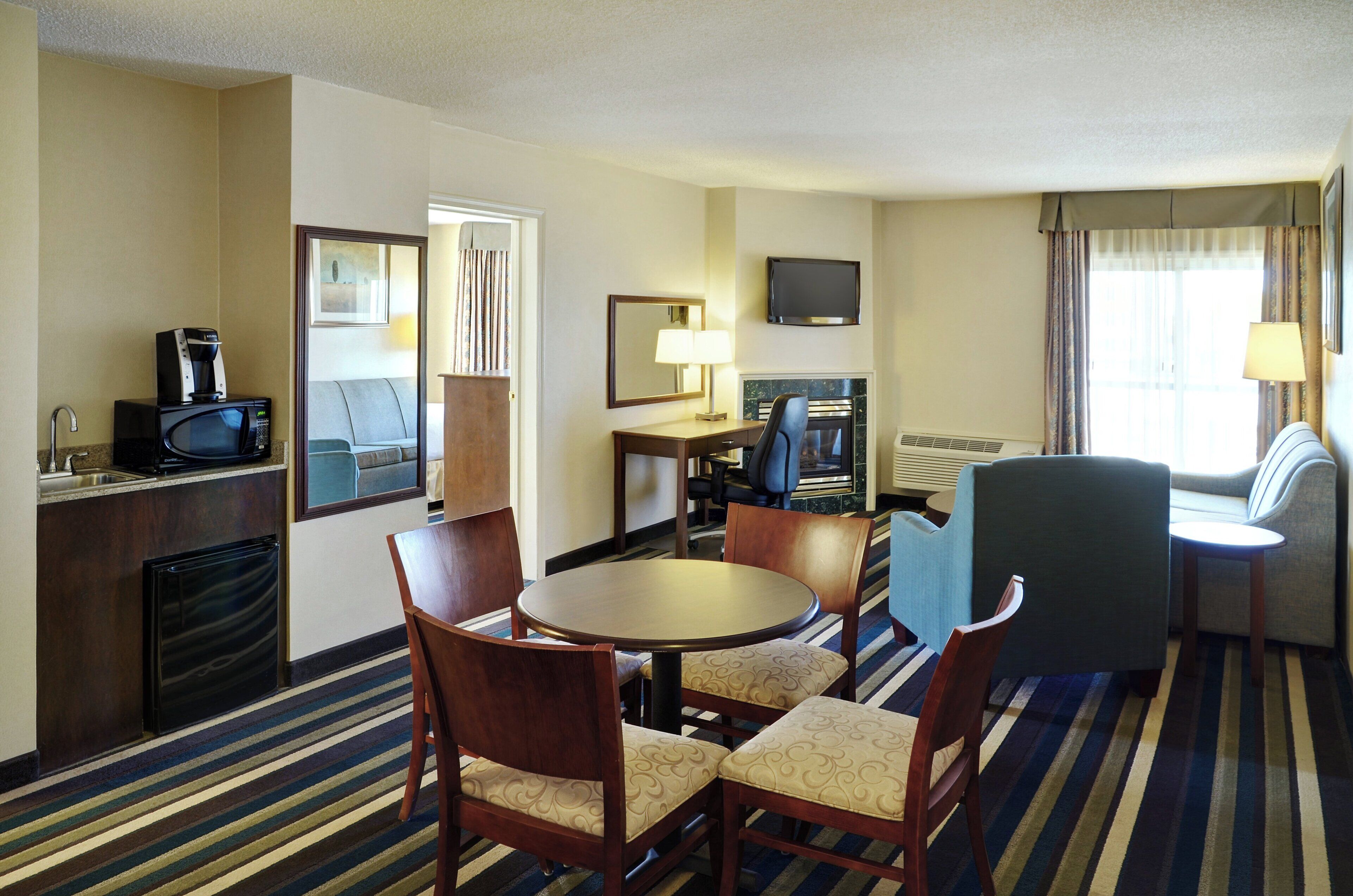 A hotel room with a dining table, chairs, a TV, a fireplace, a sofa, and a window with curtains.