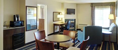 Habitación de hotel con mesa de comedor, sillas, televisor, chimenea, sofá y ventana con cortinas.