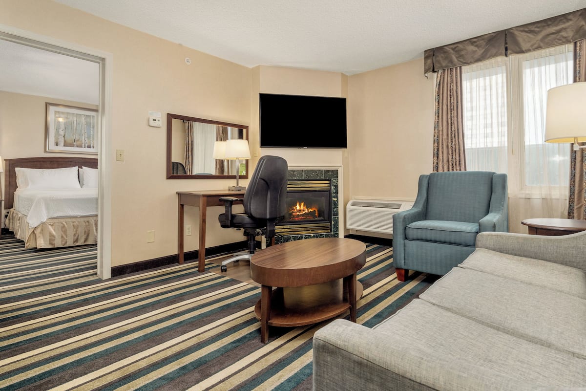 A hotel room with a bed, a desk with a chair, a television, a fireplace, a sofa, and a window with curtains.