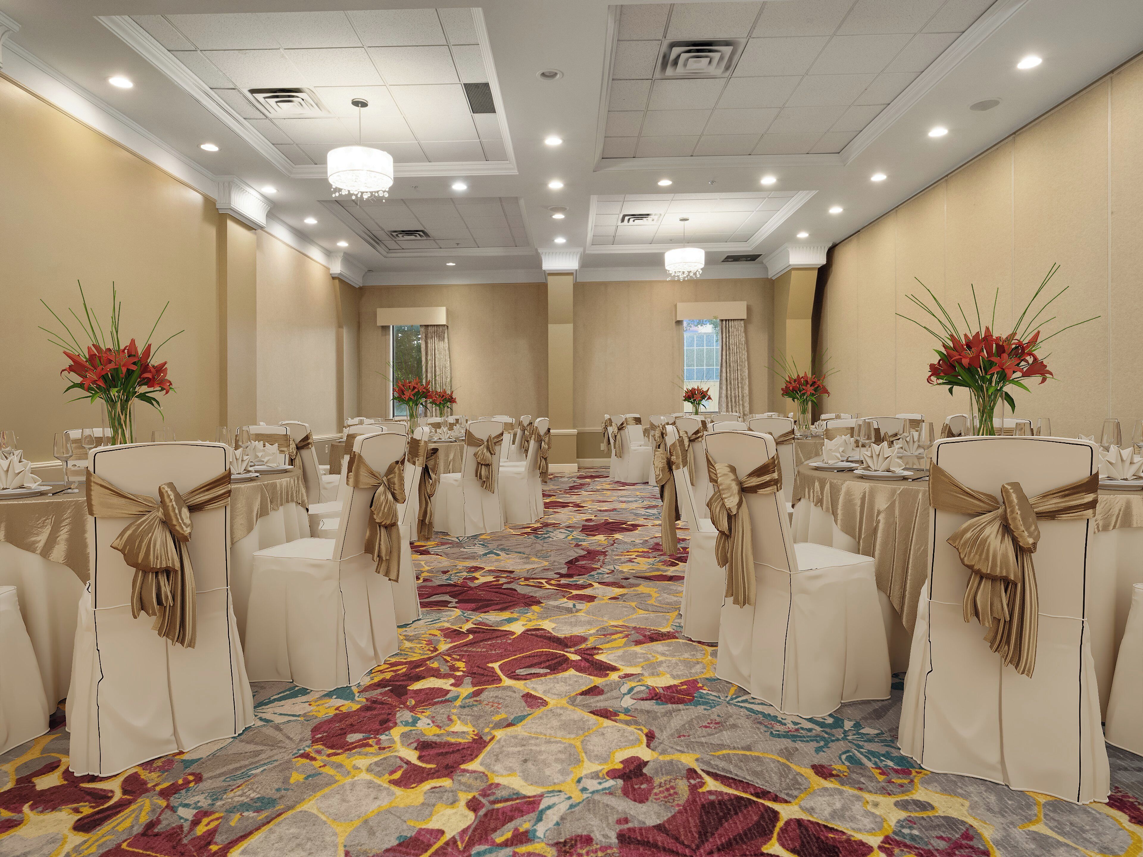 A banquet hall with rows of white chairs, gold sashes, and floral centerpieces.