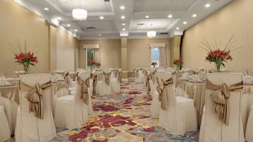 A banquet hall with rows of white chairs, gold sashes, and floral centerpieces.
