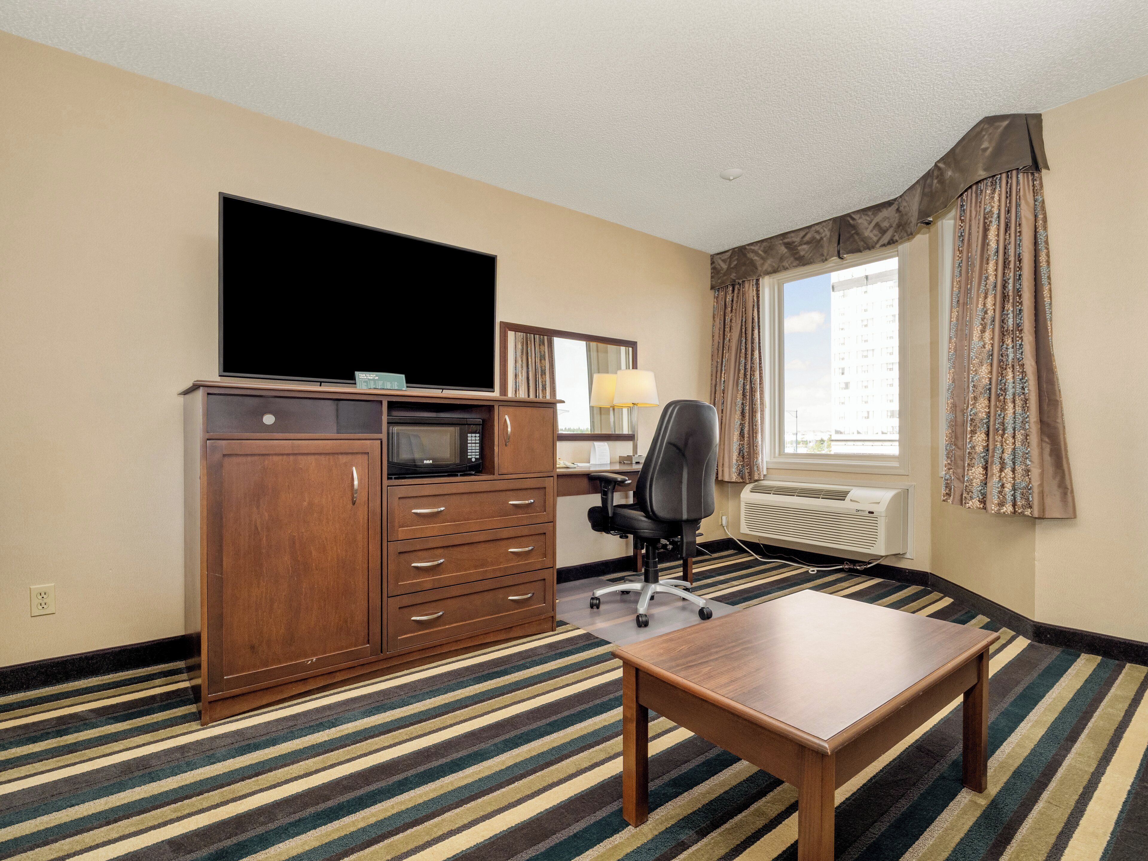 Habitación de hotel con televisor de pantalla plana, un escritorio de madera, una silla, microondas, lámpara, ventana con cortinas y alfombra a rayas.