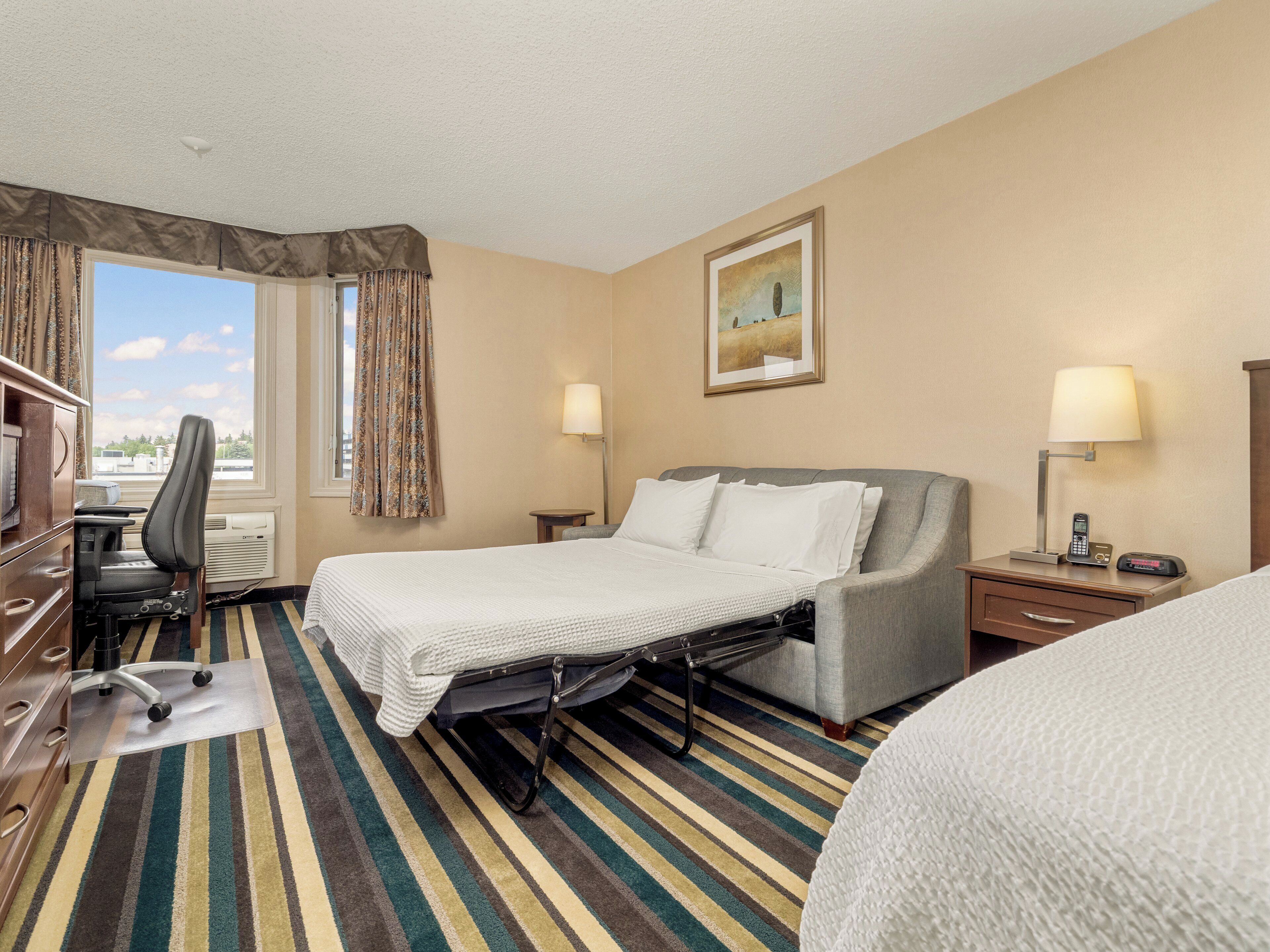 A hotel room with a bed, a desk, a chair, a lamp, and a painting on the wall.