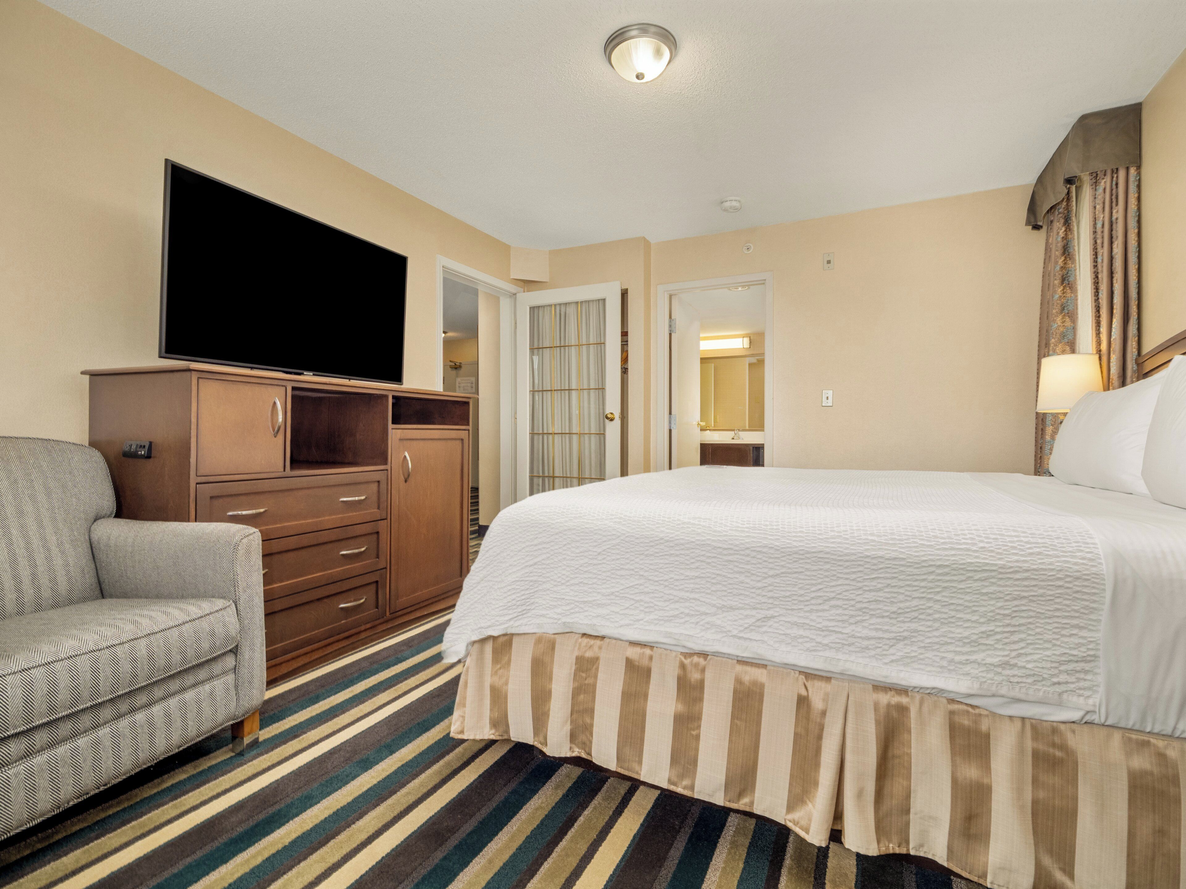 A hotel room with a large bed, a television on a wooden cabinet, a striped rug, and a chair.