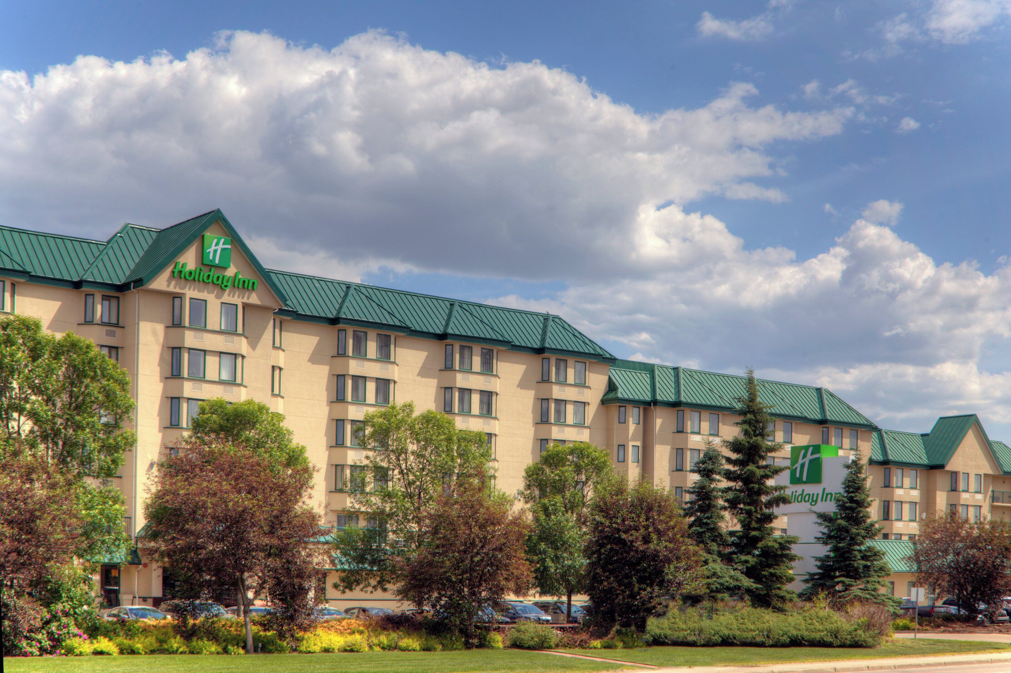 A Holiday Inn hotel with a green roof, multiple floors, and surrounded by trees and parked cars.