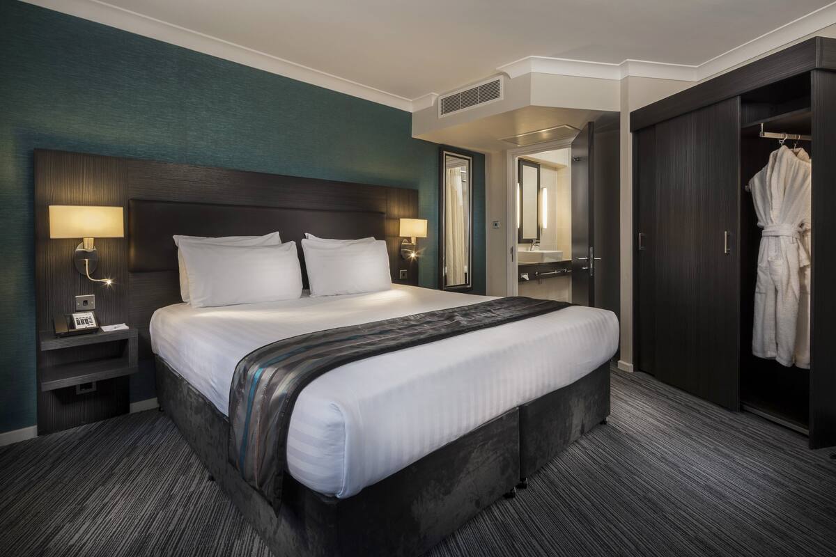 A hotel room with a large bed, two bedside lamps, a dark wood headboard, a robe hanging on a door, and a mirror above the bed.