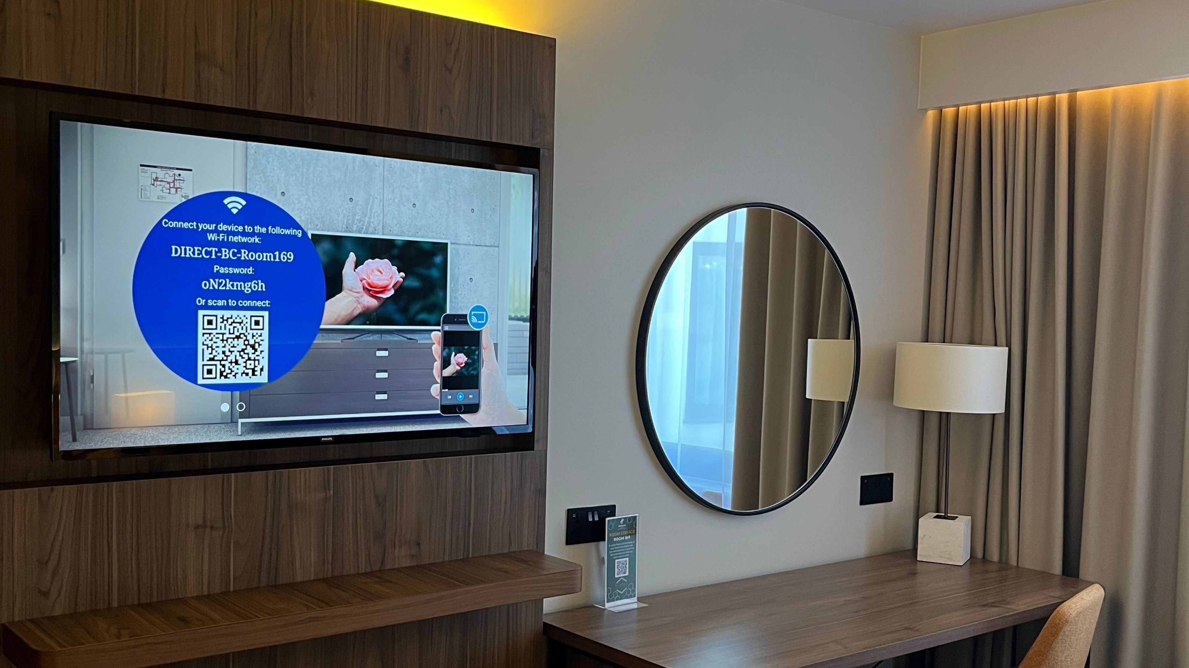 A hotel room with a mounted TV displaying a QR code for Wi-Fi connection, a round mirror, a bedside table, and a lamp.