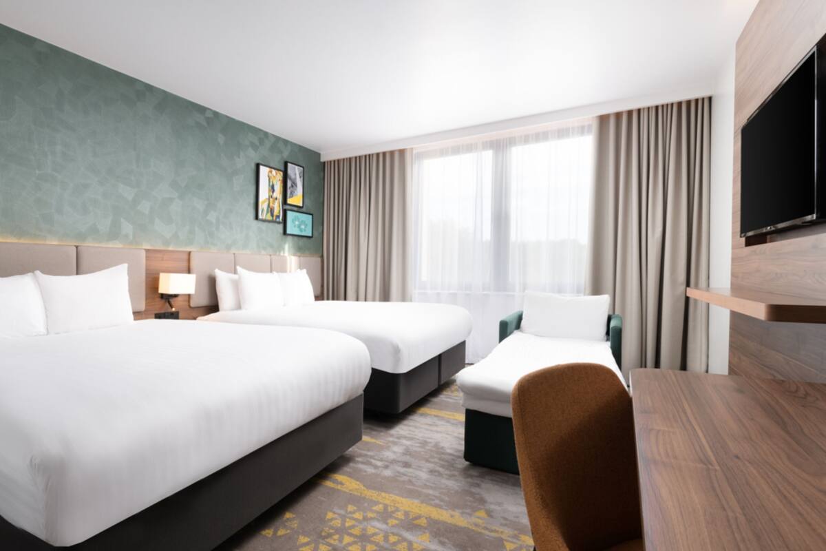 A hotel room with two beds, a desk, a TV, and a large window with curtains.