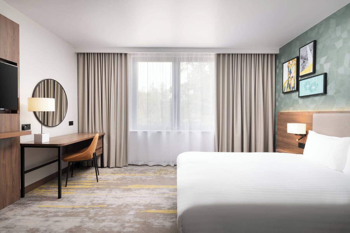 A hotel room with a bed, a desk with a chair, a TV, and a window with curtains.