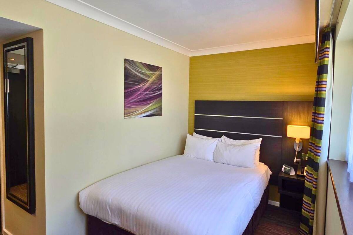 A hotel room with a large bed, a nightstand with a lamp, a painting on the wall, and a door to another room.