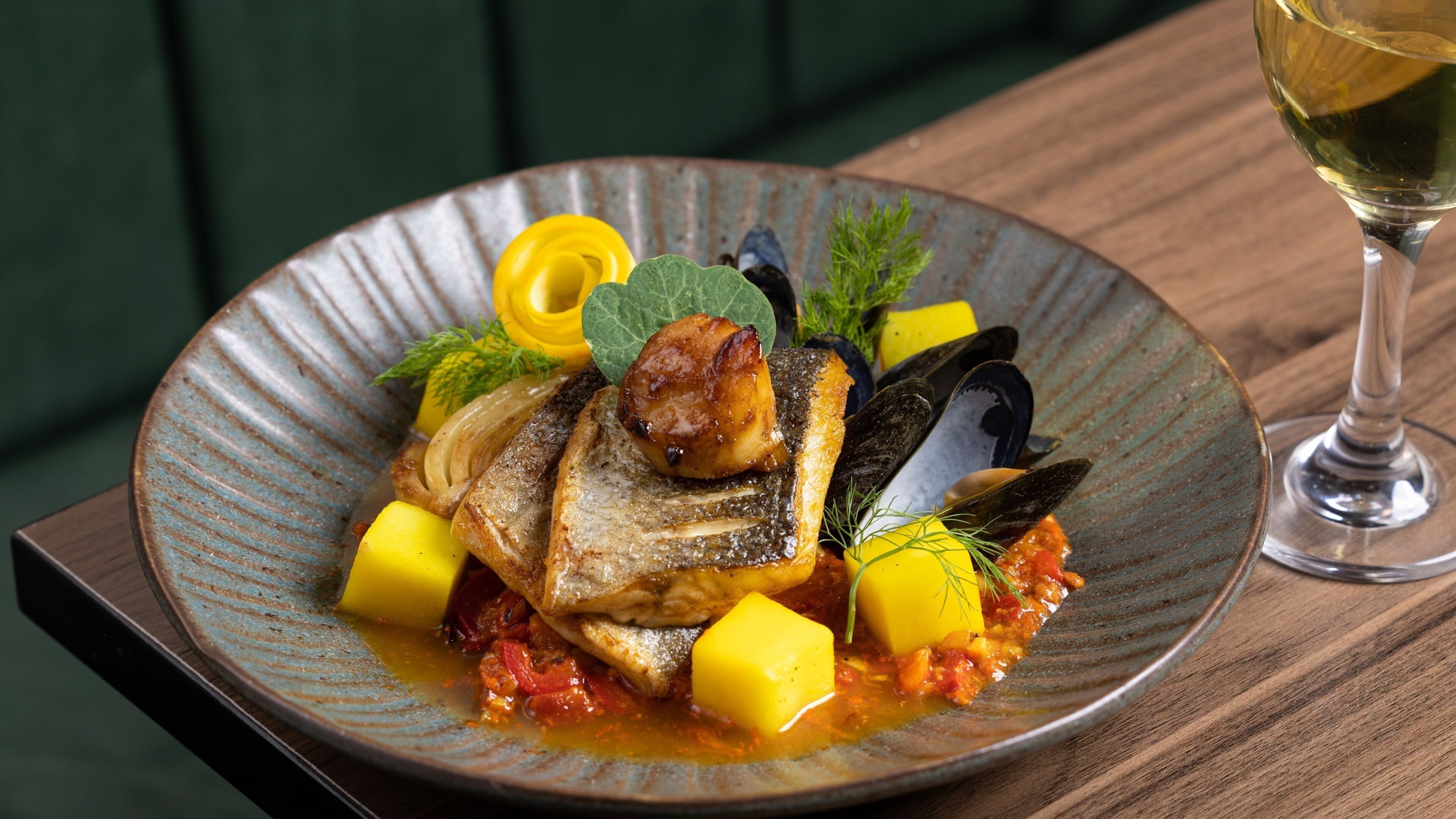 A plate of food with mussels, fish, and vegetables, accompanied by a glass of white wine on a wooden table.