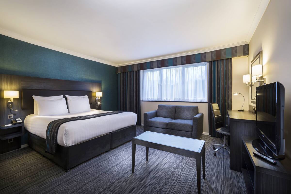 A hotel room with a large bed, a sofa, a desk with a TV, and a window with curtains.