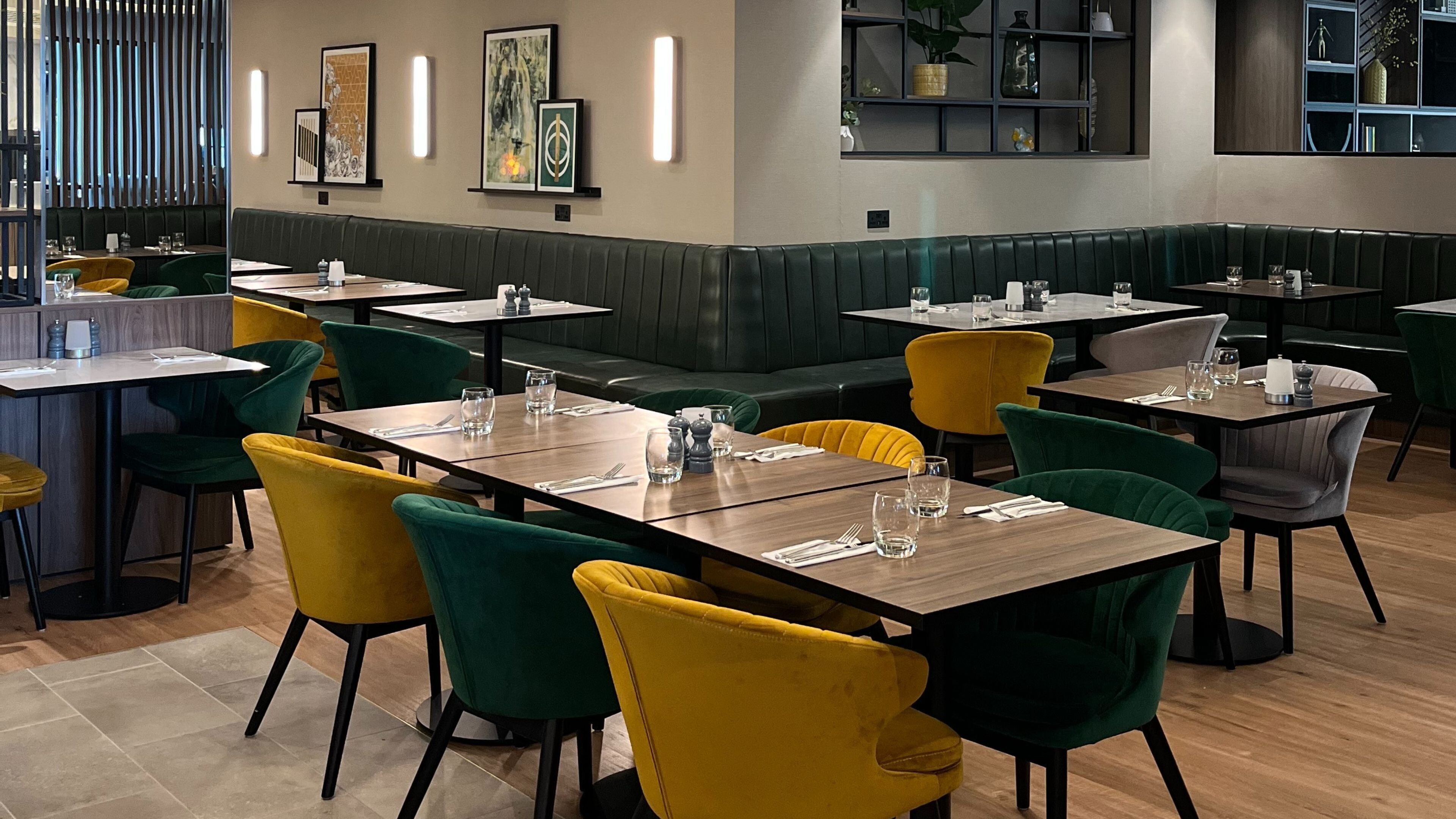 A restaurant with booths and tables, some with yellow and green chairs. There are framed pictures on the wall and a shelf with decorative items.
