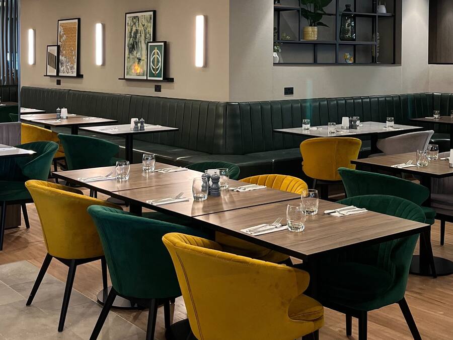 Un restaurante con cabinas y mesas, algunas con sillas amarillas y verdes. Hay cuadros enmarcados en la pared y una estantería con objetos decorativos.