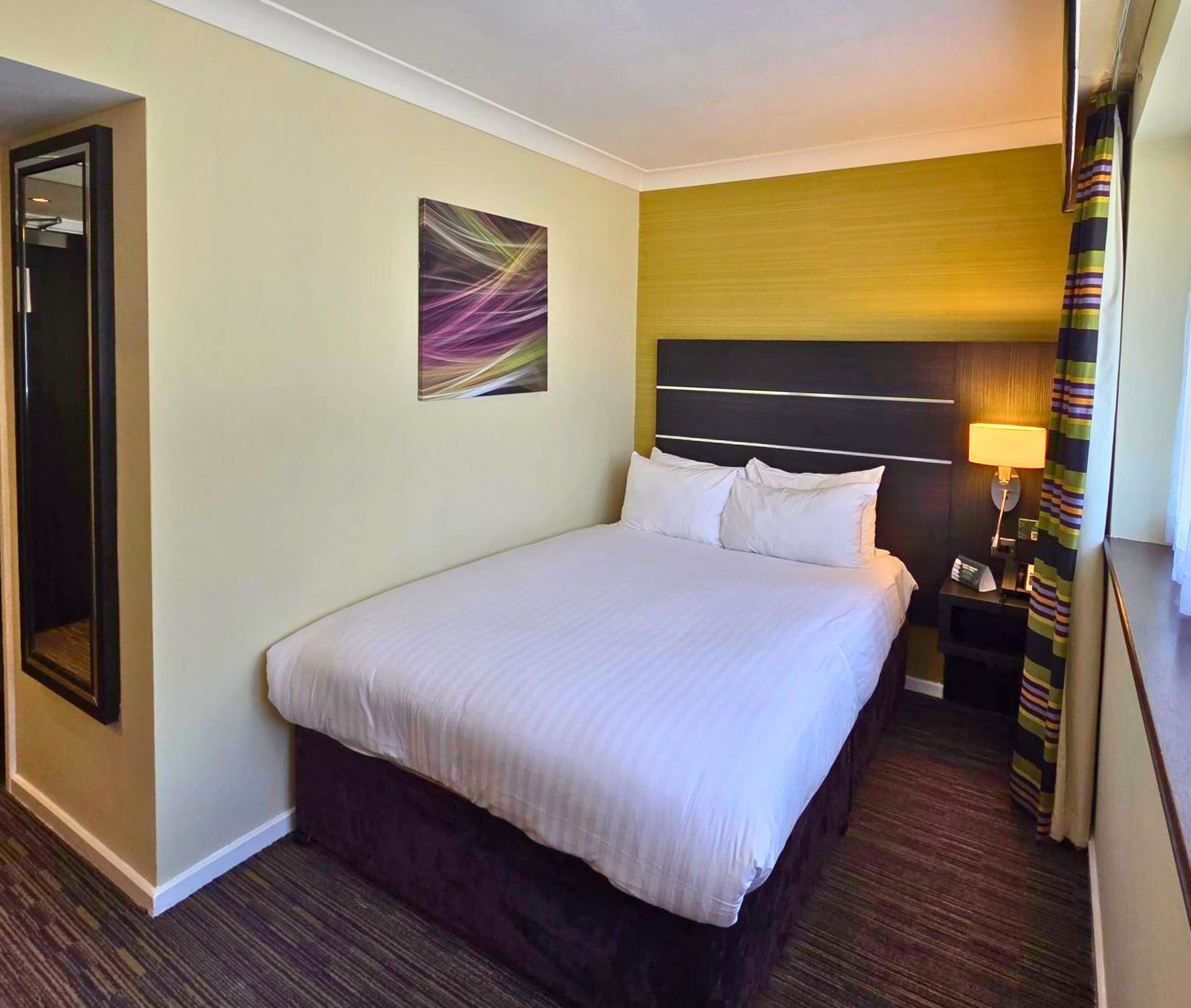 Une chambre d’hôtel avec un grand lit, une table de chevet avec une lampe, un tableau au mur et une porte donnant sur une autre pièce.