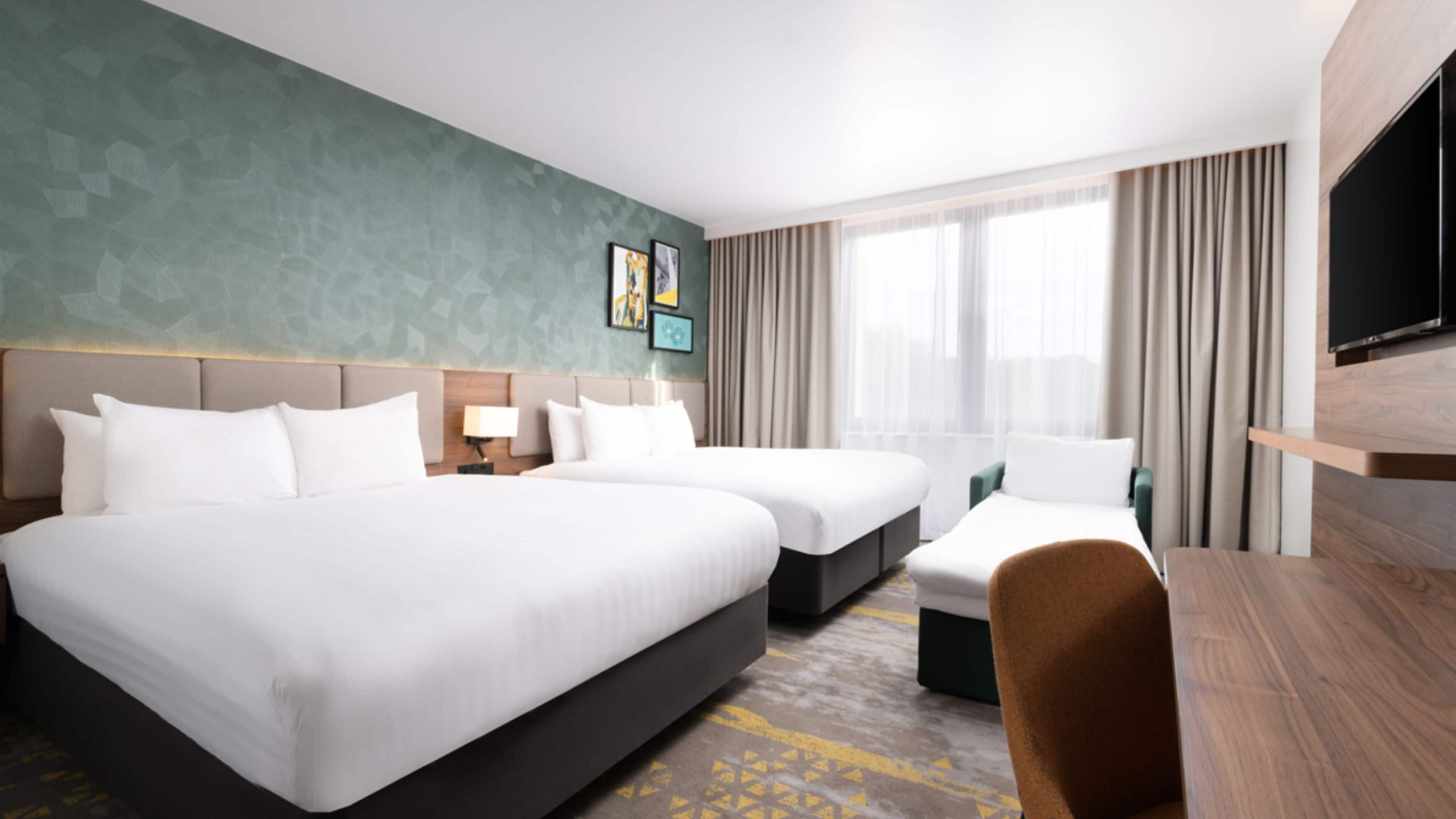 A hotel room with two beds, a desk, a TV, and a large window with curtains.
