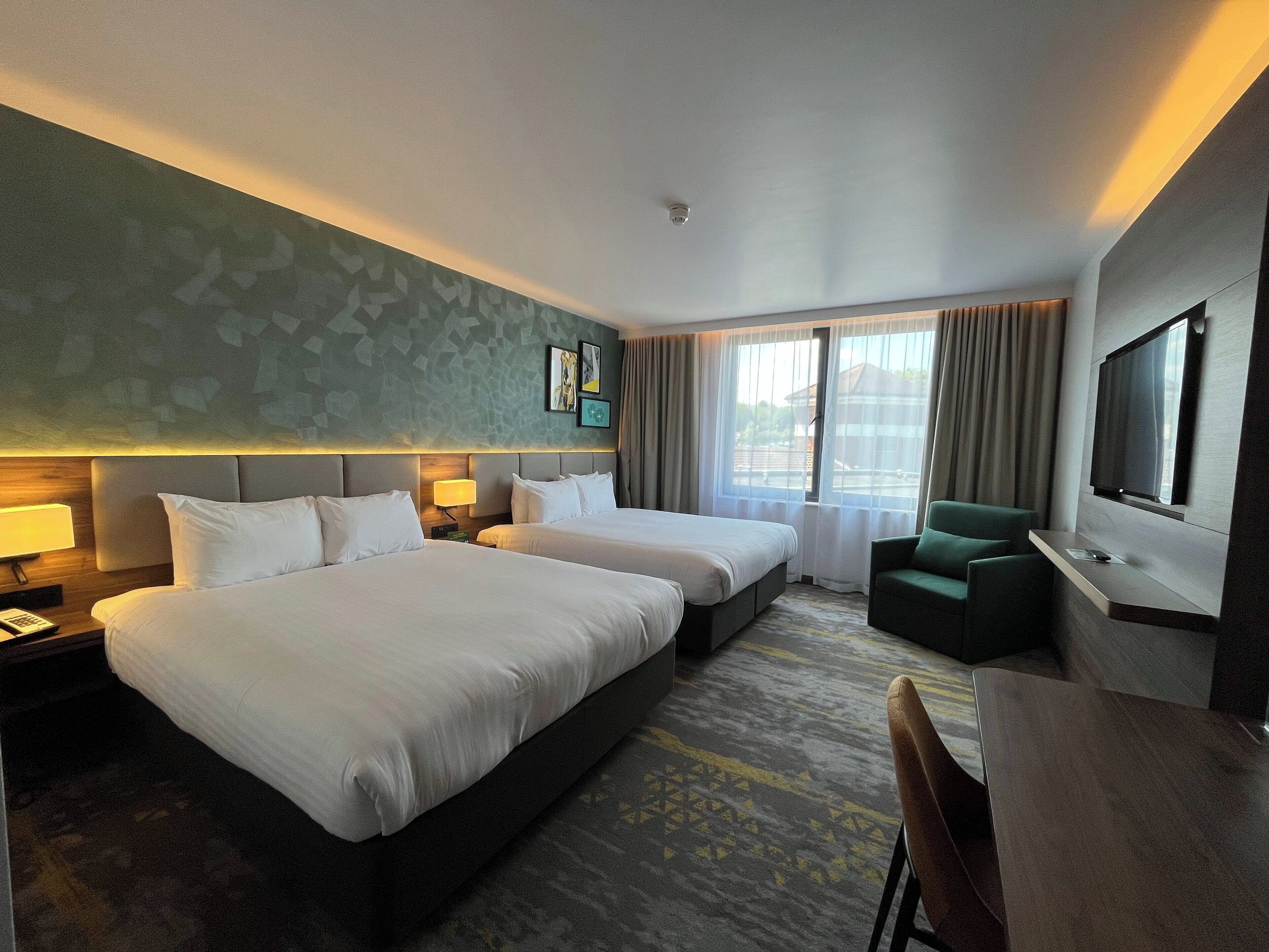 A hotel room with two beds, a desk with a chair, a TV, and a window with curtains.