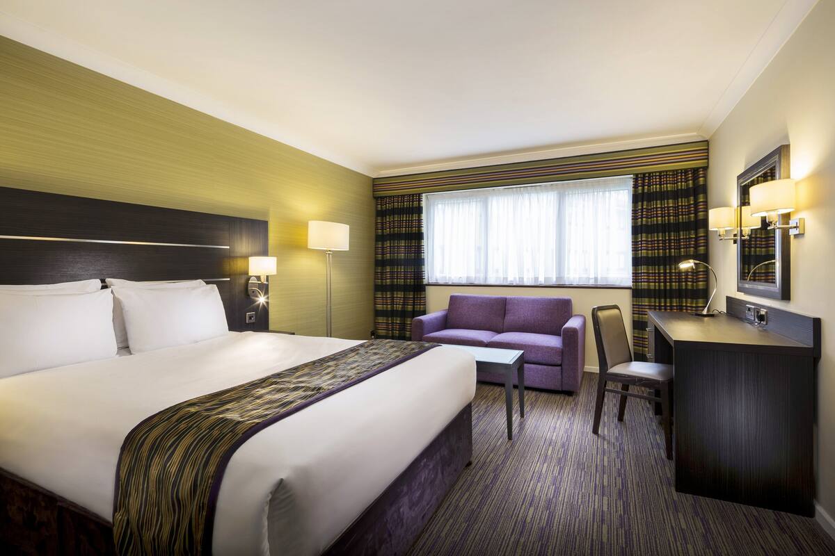 A hotel room with a large bed, a desk, a chair, a sofa, and a window with curtains.