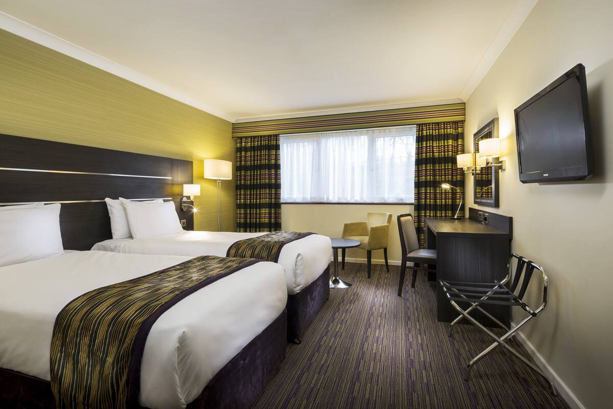 A hotel room with two beds, a desk, a chair, a TV, and a window with curtains.