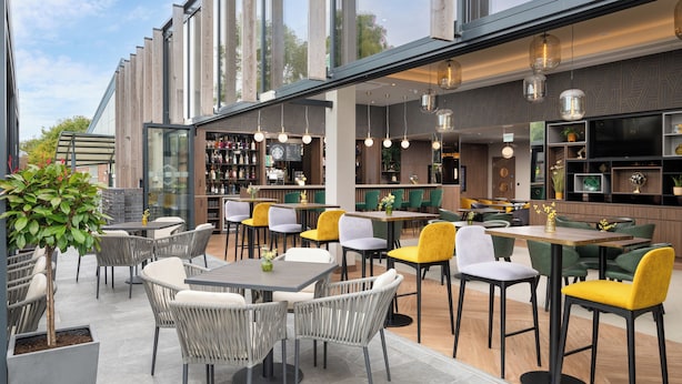 An outdoor dining area with multiple tables and chairs, a bar area with shelves and a television, and large windows providing views of the surroundings.