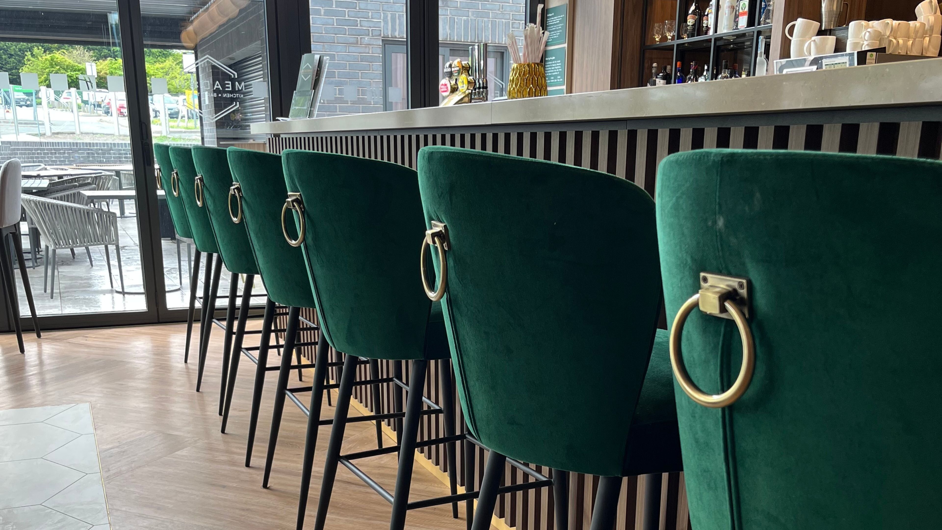 A row of green upholstered bar stools with metal rings, a bar area with various items, and a view of a parking lot through large windows.