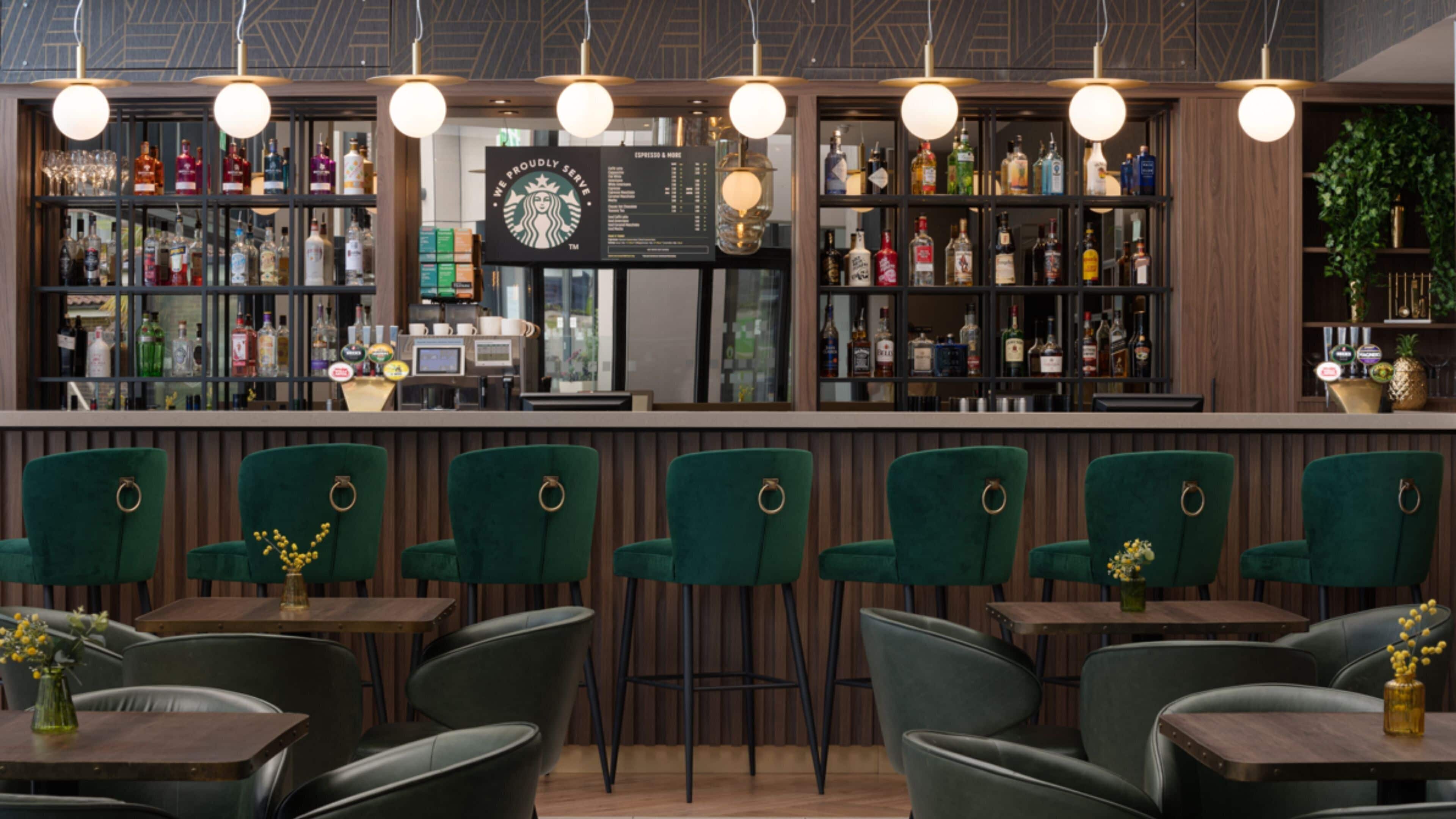 Zona de bar con taburetes verdes tapizados, una variedad de botellas de licor y un panel con la carta.