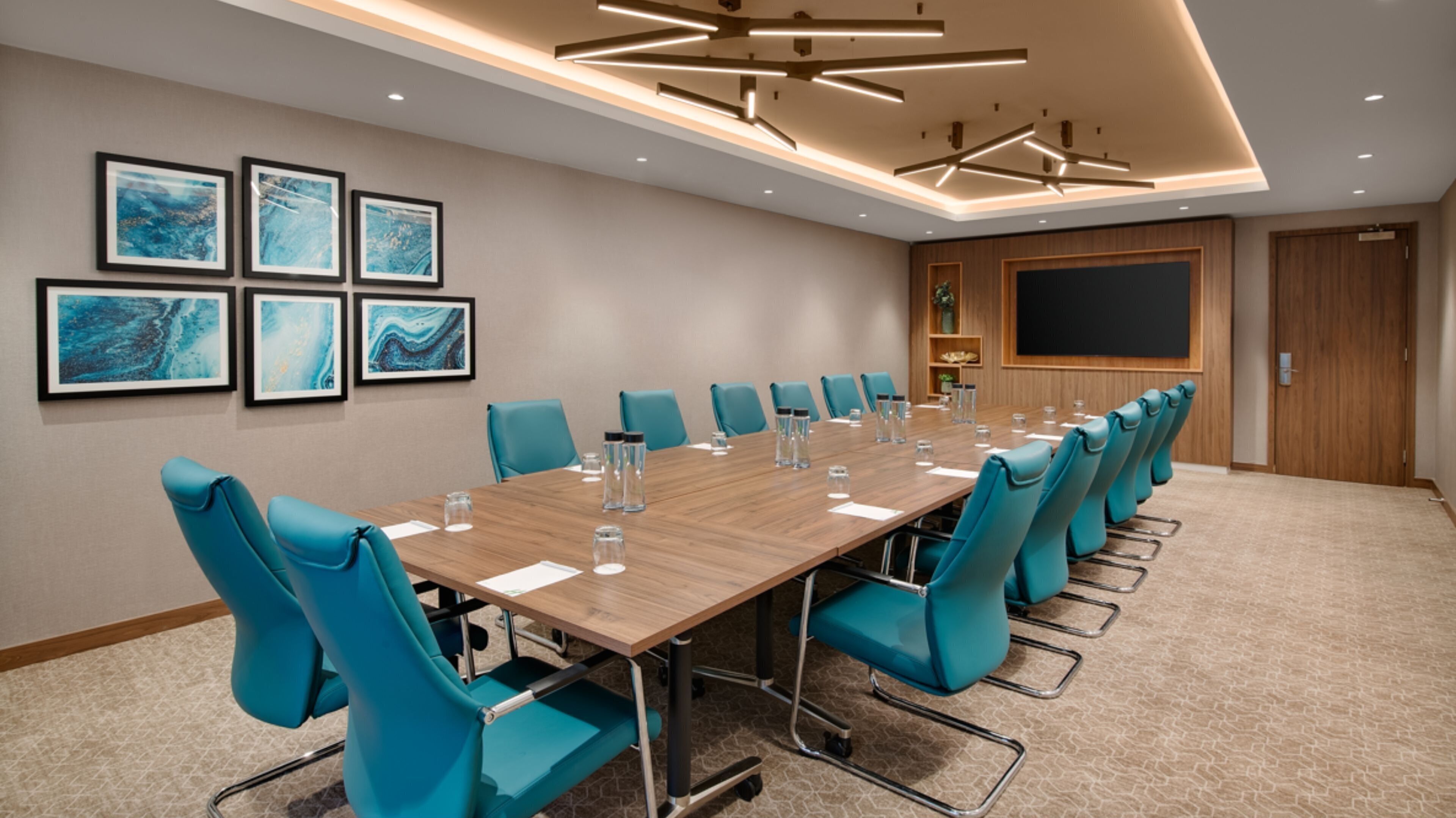 A conference room with a long table, teal chairs, framed artwork on the wall, and a projector screen.