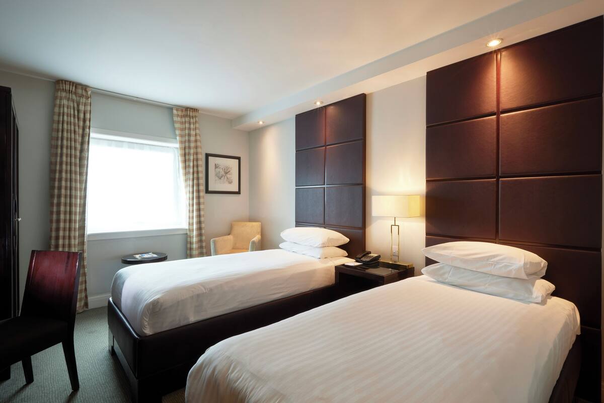 A hotel room with two beds, a desk, a chair, a bedside table with a lamp, and a window with curtains.