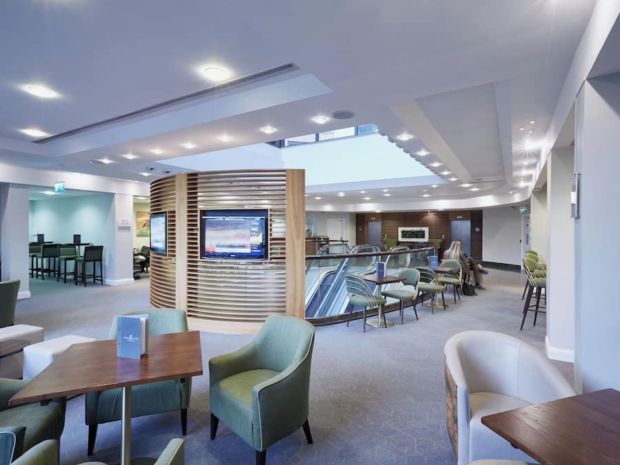 A modern hotel lobby with seating areas, a reception desk, a TV screen, and an escalator.