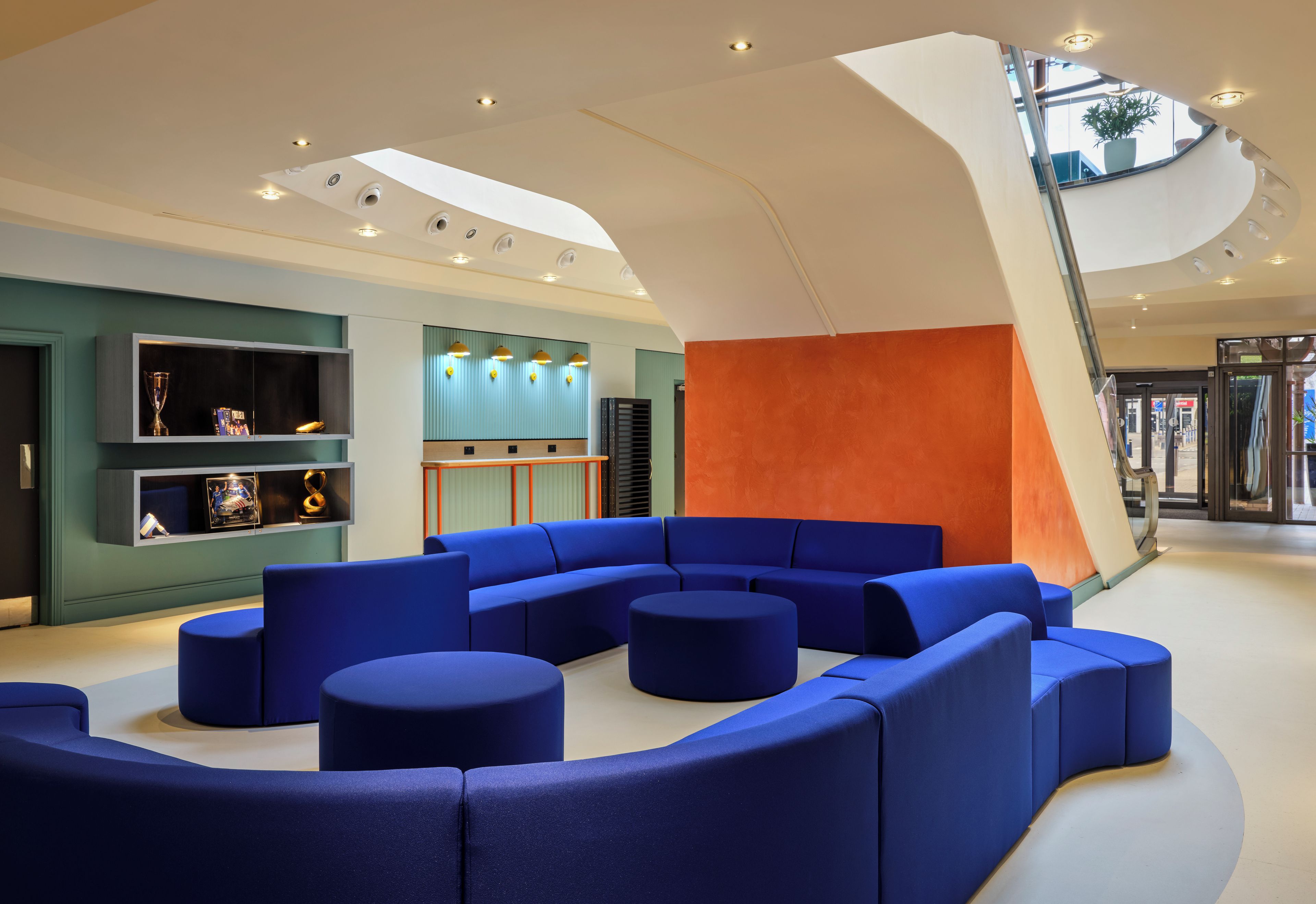 Un hall d’entrée moderne avec un espace détente bleu aux lignes courbes, un escalier en verre et un mur agrémenté d’un luminaire vertical.