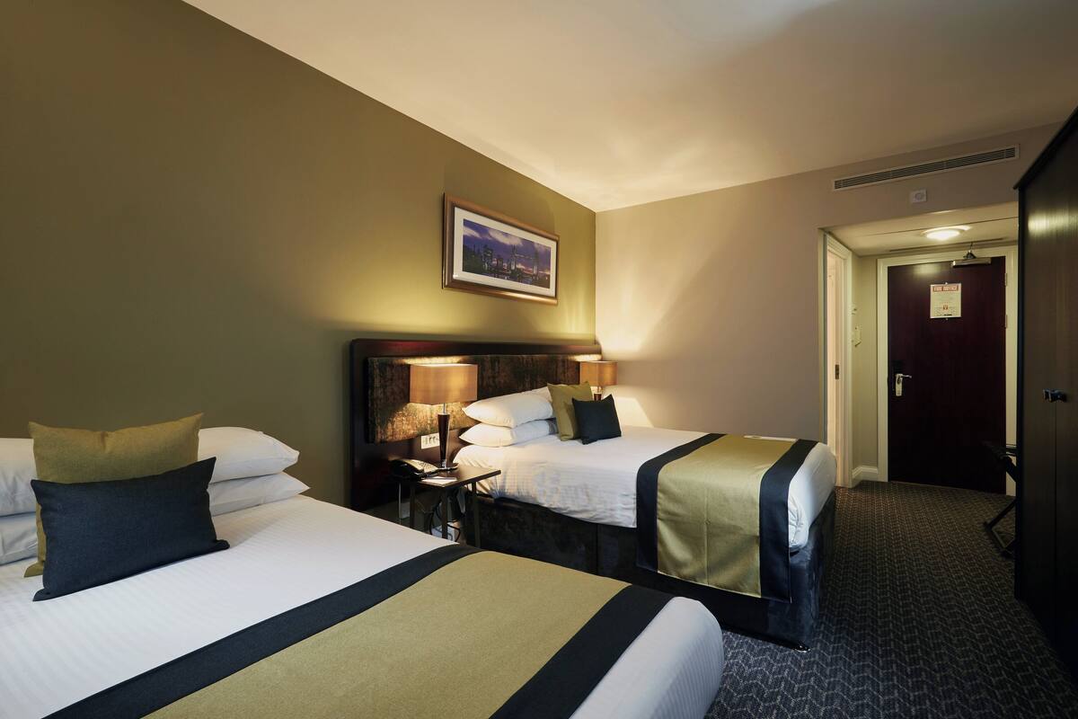 A hotel room with two beds, a nightstand with a lamp, a framed picture on the wall, and a door to the right.