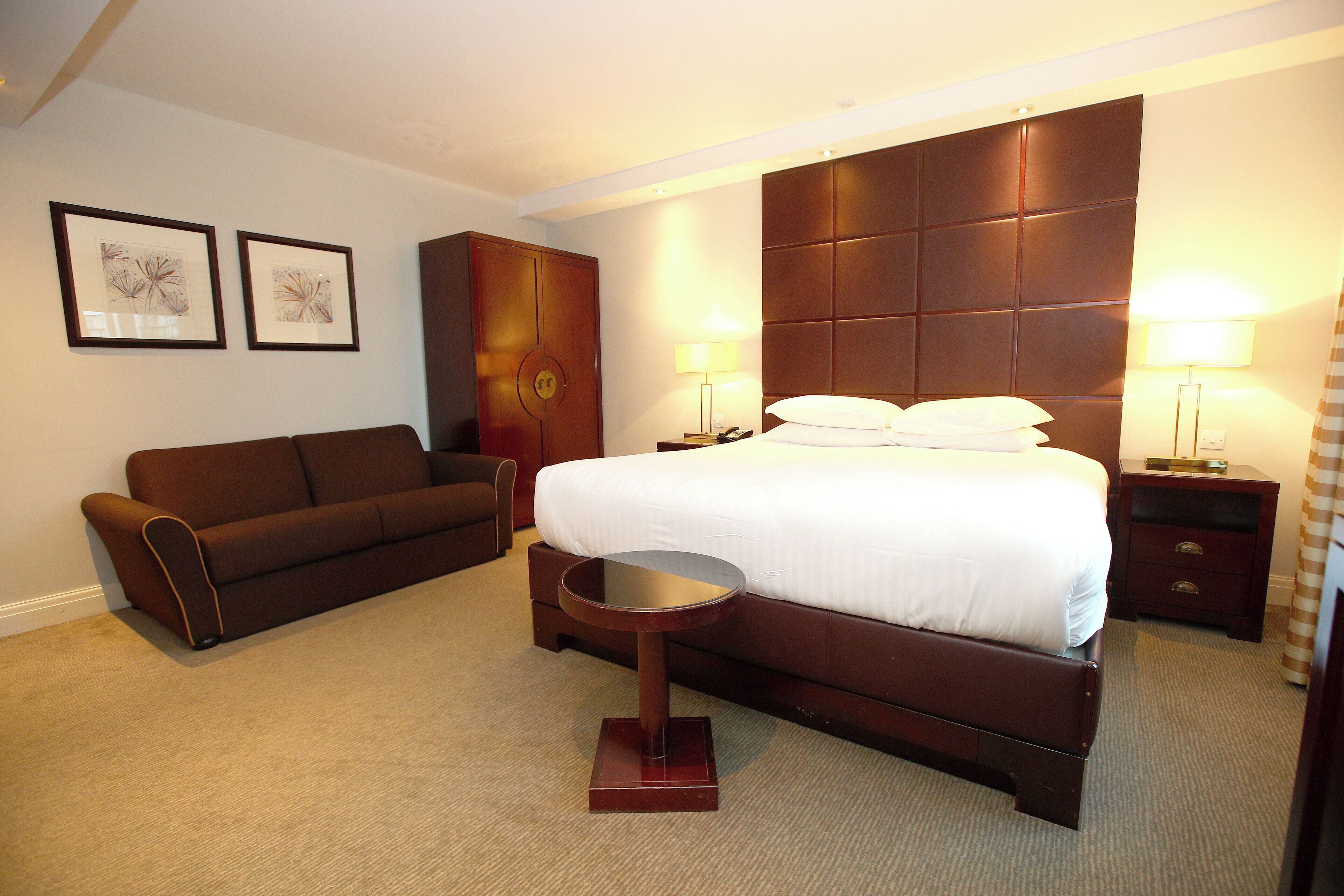 A hotel room with a large bed, a bedside table, a nightstand, a round side table, a sofa, and two framed pictures on the wall.