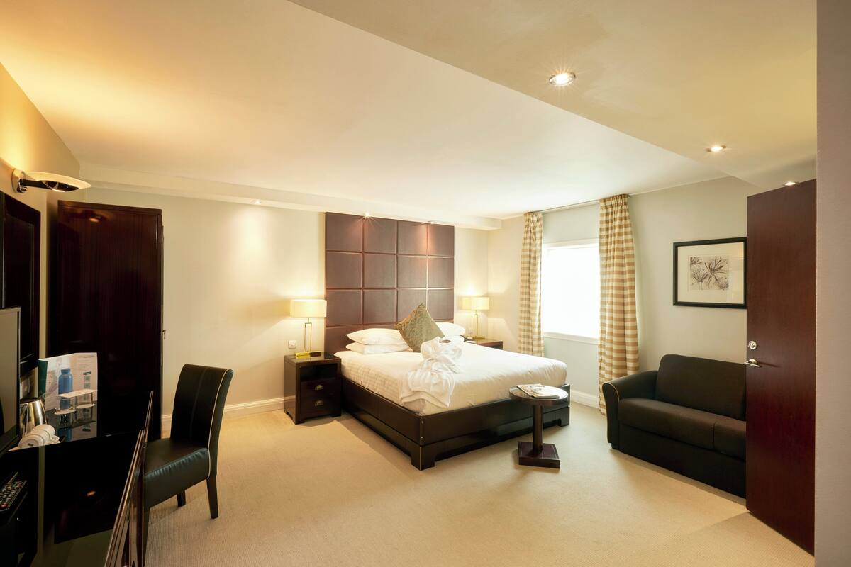 A hotel room with a large bed, bedside tables with lamps, a desk with a chair, a sofa, and a wardrobe.