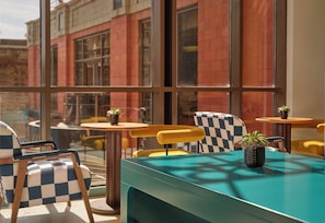 An outdoor seating area with checkered chairs and tables, a teal table, and a view of a red brick building through large windows. - lyf Chelsea London (London)