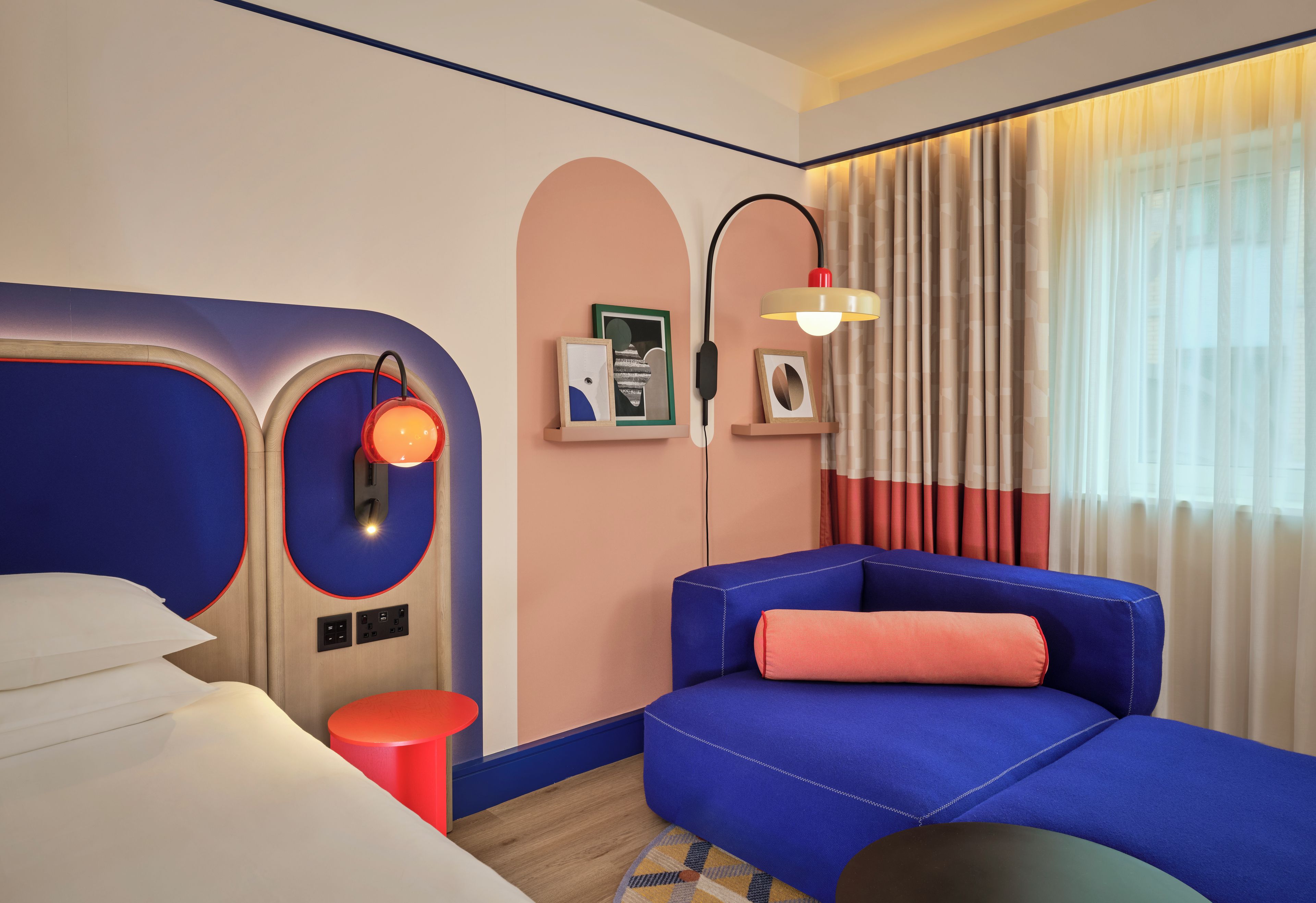 Habitación de hotel con cama, zona de estar con sofá azul y cojín rojo, una mesita pequeña roja, una lámpara de pared y ventana con cortinas.