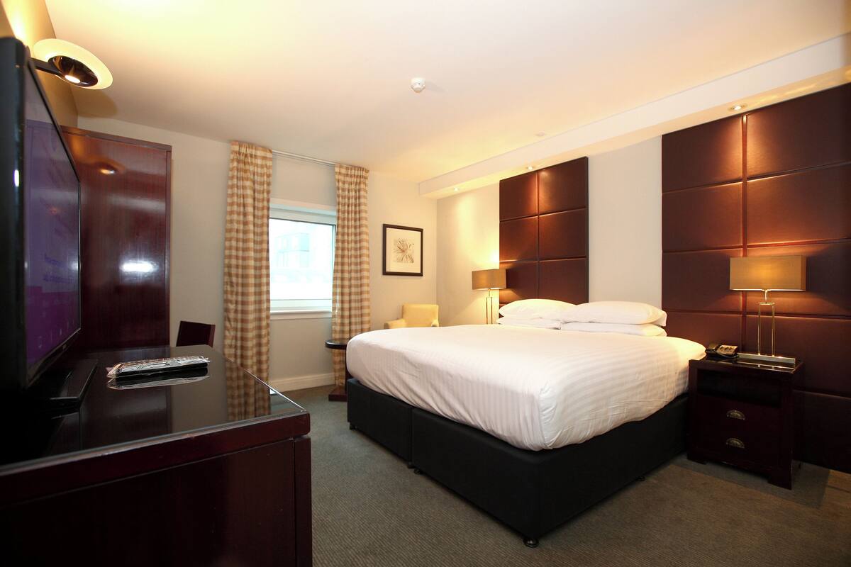 A hotel room with a bed, bedside table, lamp, and a TV on a desk.