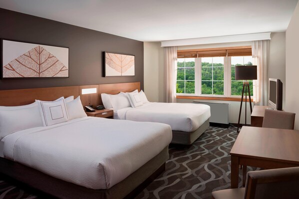 Une chambre d’hôtel avec deux lits, un bureau, une chaise, une lampe et une fenêtre donnant sur un paysage verdoyant.