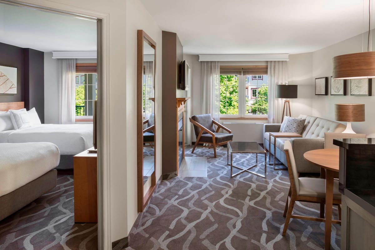 Ein Hotelzimmer mit einem Bett, einem Schreibtisch, einem Stuhl und einer Couch. Es gibt Fenster mit Vorhängen, eine Lampe und einen Fernseher.