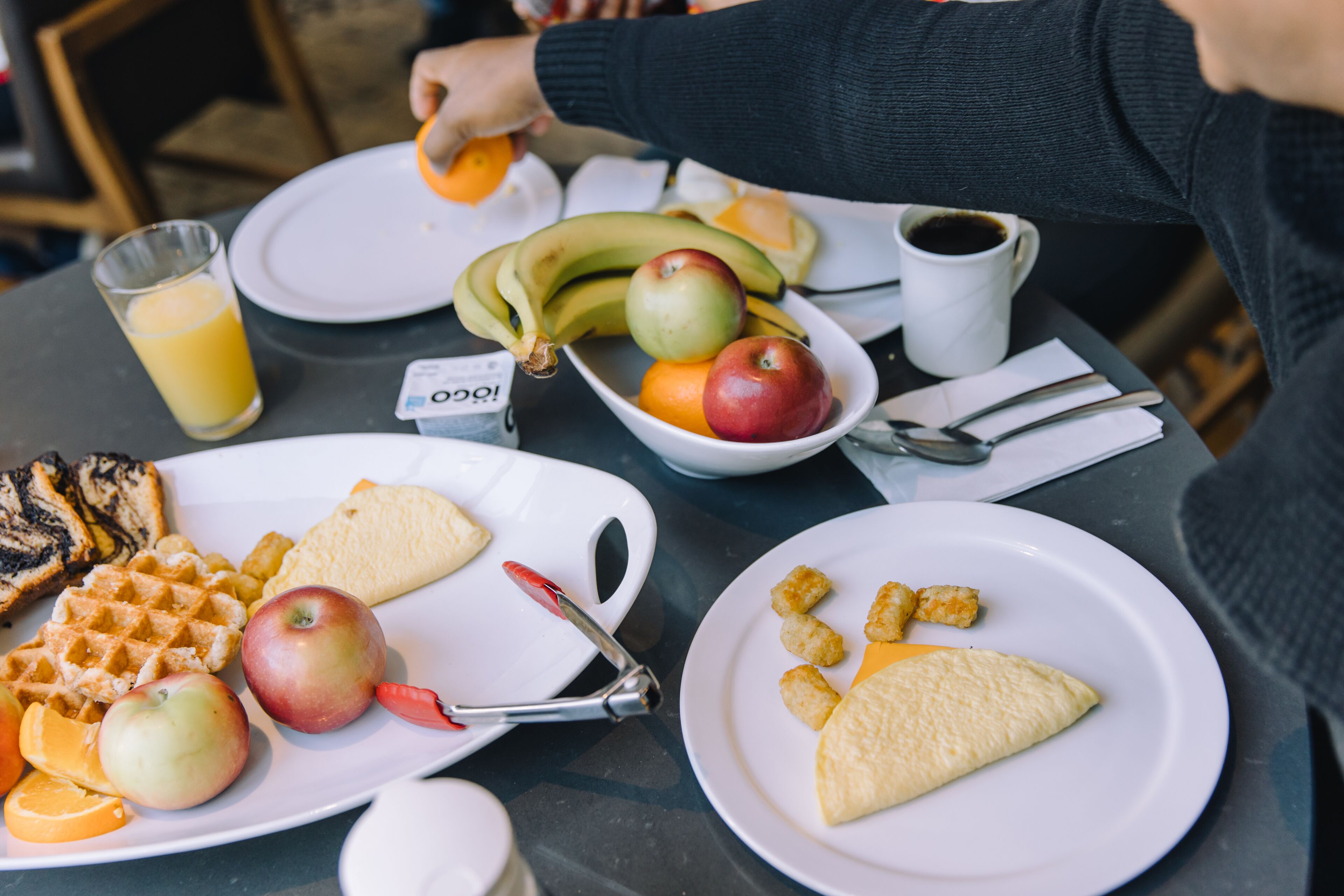 A breakfast table with plates of waffles, apples, bananas, and orange juice.