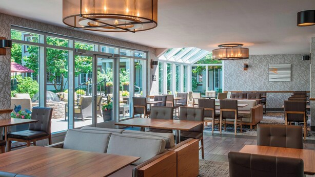 A modern restaurant with a mix of booths and tables, large windows offering a view of the outdoor garden, and contemporary lighting fixtures.