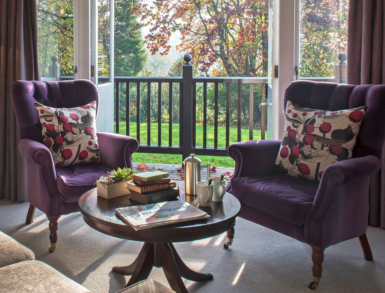 Een kamer met twee paarse fauteuils, elk met een bloemenkussen, een ronde bijzettafel met boeken en een theepot, en uitzicht op een groene tuin door grote ramen.