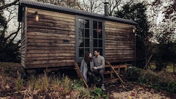 A wooden cabin with a large window and a door, situated in a natural setting with trees and foliage.