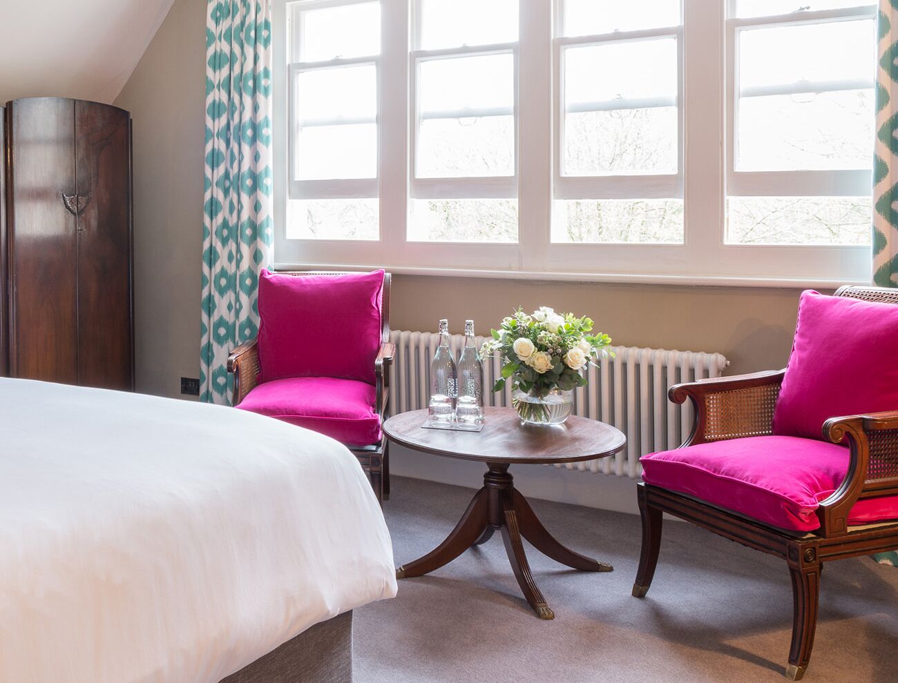 A hotel room with a bed, two chairs, a round table with a vase of flowers, and large windows.