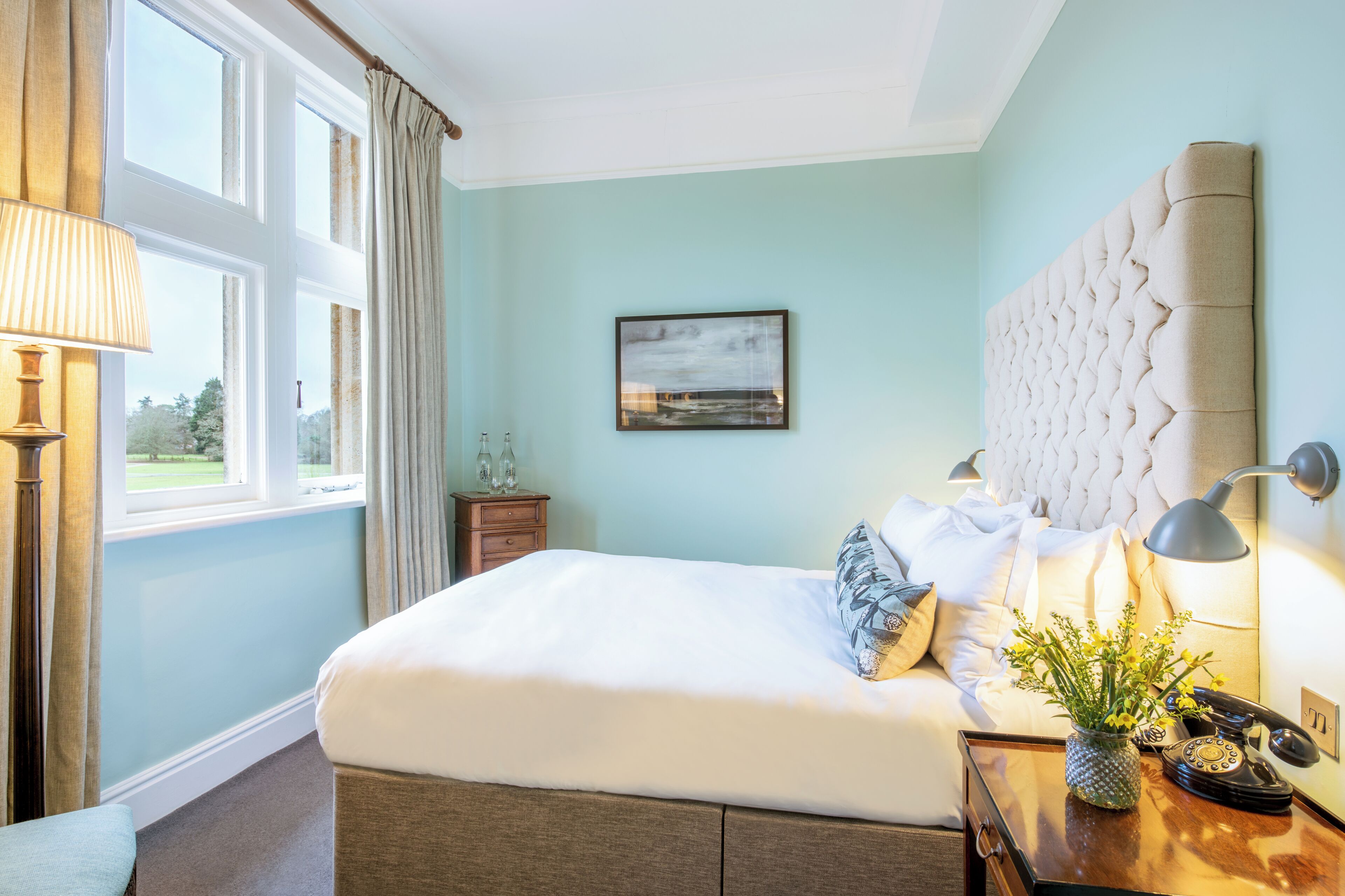 A neatly made bed with a headboard, a bedside lamp, a vase with flowers, and a telephone on a bedside table.