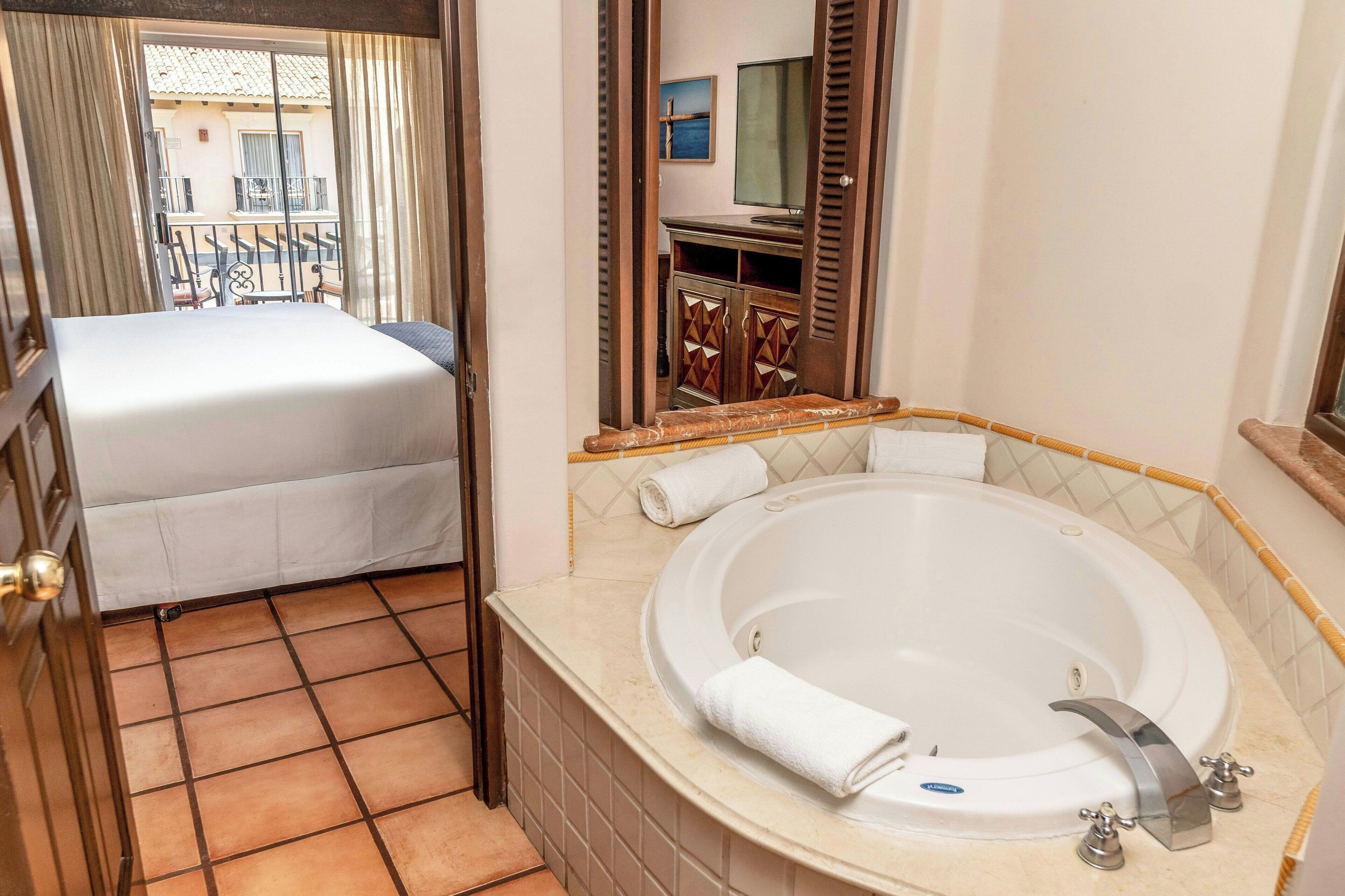 A hotel room with a bed, a large bathtub, and a view of the outside balcony.