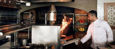A chef performing a flambé with a large metal pot on a stove in a kitchen with brick walls and a tiled backsplash.