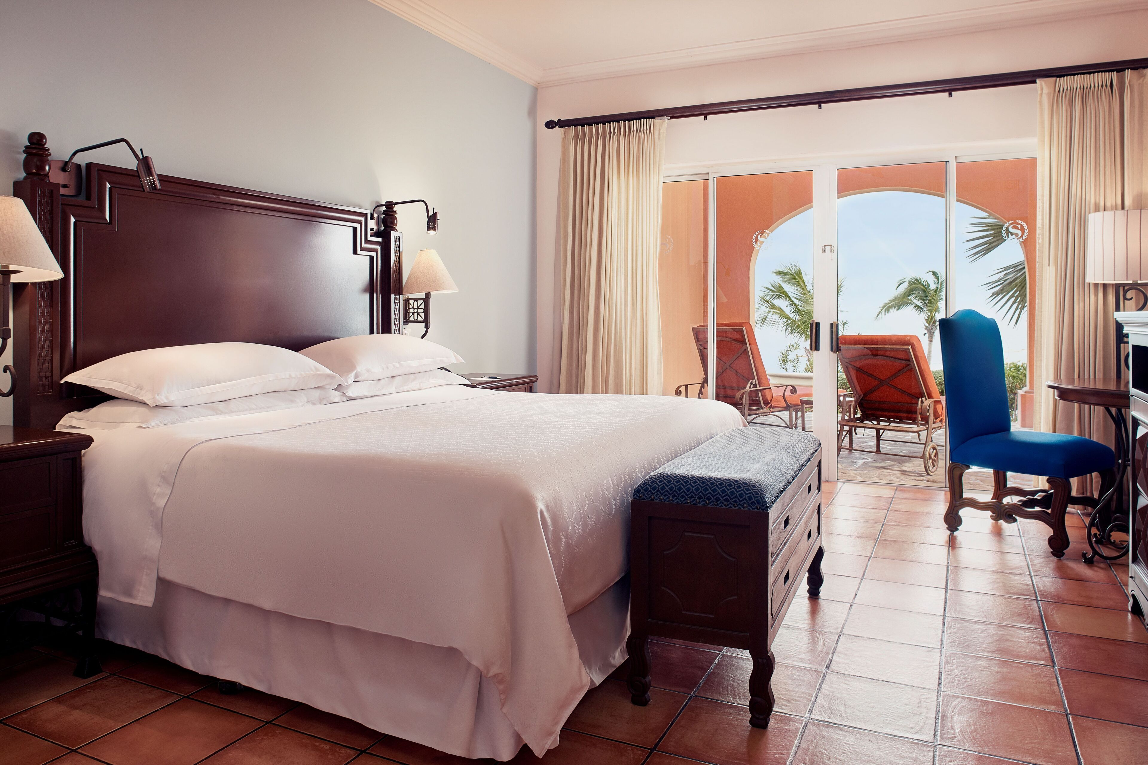 Une chambre d’hôtel avec un grand lit, un bureau avec une chaise et un balcon offrant une vue sur les palmiers.