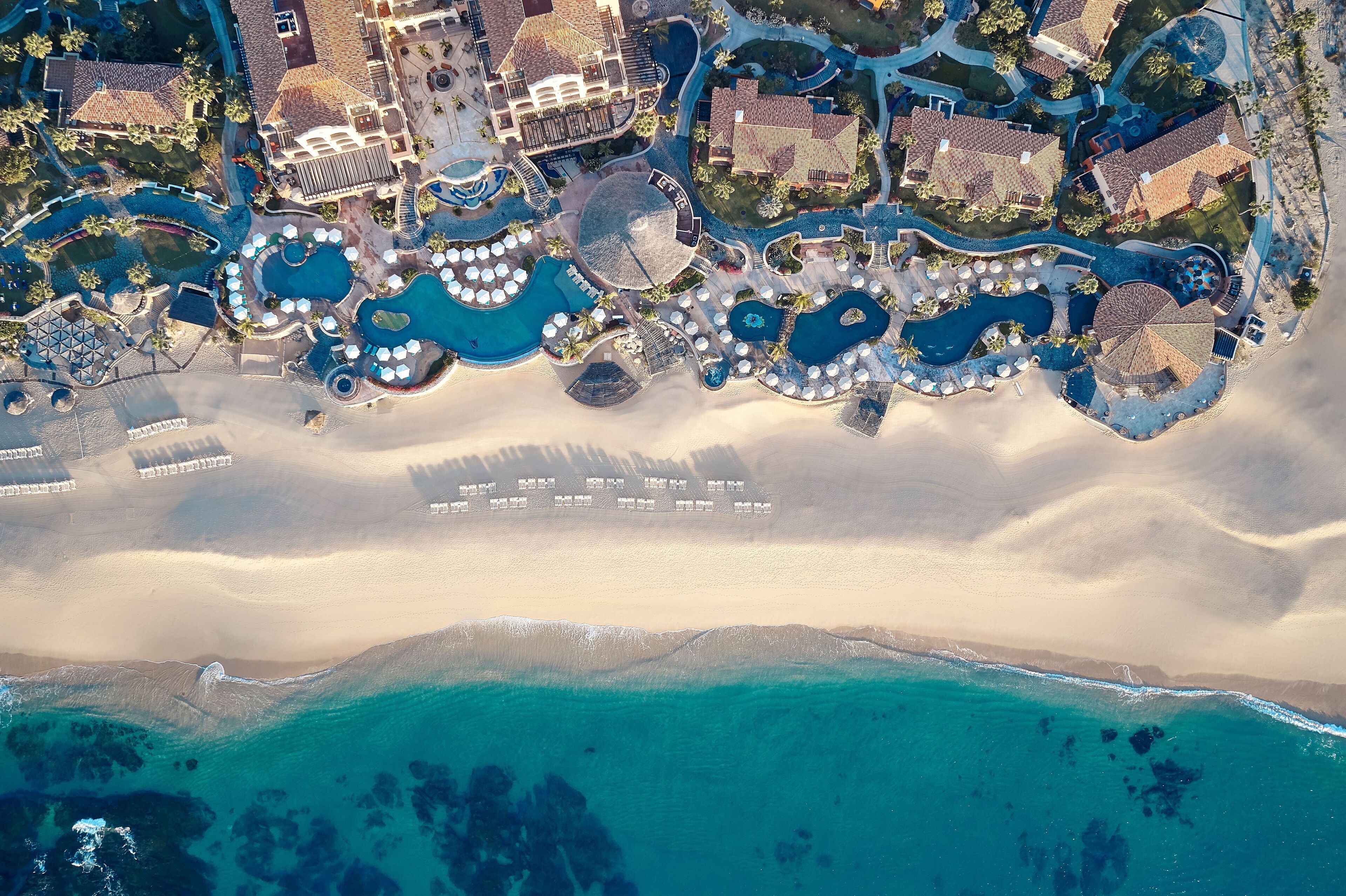 Vista aérea de un resort con múltiples piscinas, villas frente a la playa y una playa de arena.