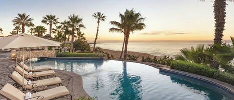 A poolside area with lounge chairs and umbrellas, palm trees, and a view of the ocean.